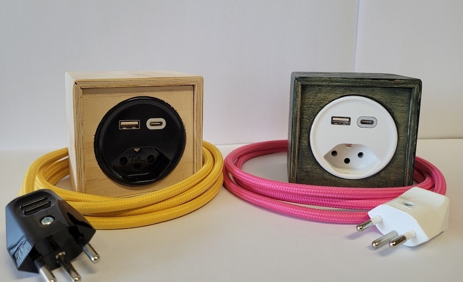 Man sieht zwei Power-Cubes. Einen hellen, mit einer schwarzen Steckdose sowie USB- und USB-C-Anschluss mit gelbem Kabel und schwarzem Stecker sowie einen zweiten, dunklen mit weisser Steckdose sowie USB- und USB-C-Anschluss mit pinkem Kabel und weissem Stecker. 